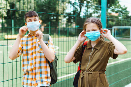 two teenagers boy and girl on the way to the school, they use protective face masks, stand by the playground and show social distance to protect against coronavirus infectionの写真素材