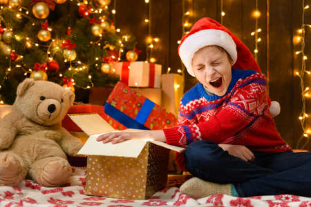 Child girl posing in new year or christmas decoration. Festive lights and lots of gifts, an elegant Christmas tree with toys. The girl is wearing a red sweater and a Santa hat, she knitsの写真素材