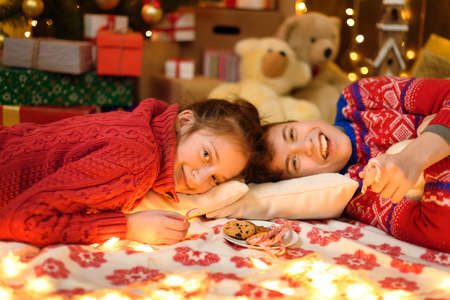 Portrait of boy and girl in new year decoration. They lie and eat sweets and cookies. Holiday lights, gifts and christmas tree decorated with toys.の写真素材