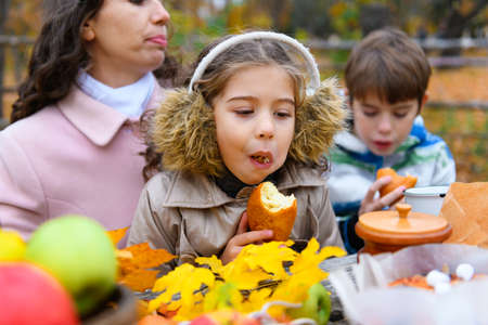 Portrait of a girl eating a bun. Happy family resting in autumn city park. People are sitting at the table, eating and talking. Posing against the background of beautiful yellow trees.の写真素材