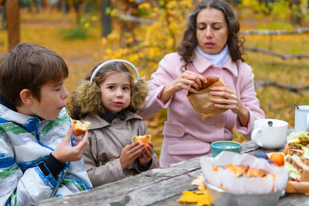 Portrait of a happy family in autumn city park. People are sitting at the table, eating and talking. Posing against the background of beautiful yellow trees. They are happy together.の写真素材