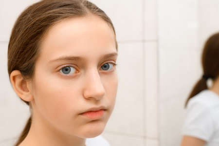 face of teenage girl, she looks at herself in the mirror and touches her face with her hand, she is upset and has frustrated emotionsの写真素材