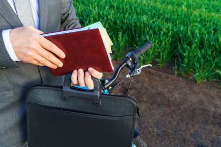 business concept - A businessman rides a bicycle on a green grass field, he takes a diary out of his briefcase and reads, dressed in a business suitの写真素材