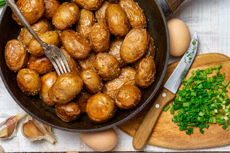fried potatoes in a frying pan, garlic and green onions, farm eggs and salt, raw and cooked food in a rustic style on a background of white wooden boardsの写真素材