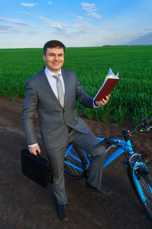 business concept - A businessman rides a bicycle on a green grass field, looks into the distance through binoculars, dressed in a business suit, he has a briefcase and documentsの写真素材