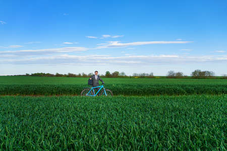 business concept - A businessman rides a bicycle on a green grass field, looks into the distance through binoculars, dressed in a business suit, he has a briefcase and documentsの写真素材