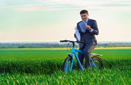 business concept - A businessman rides a bicycle on a green grass field, looks into the distance through binoculars, dressed in a business suit, he has a briefcase and documentsの写真素材