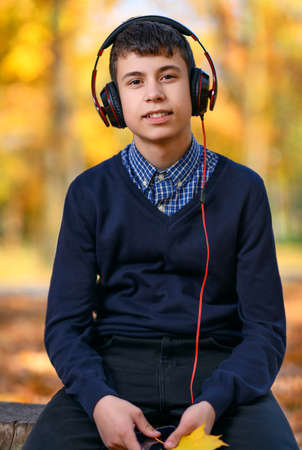 a teenage boy sits in autumn park and listens to music with headphones, trees with yellow leaves on...