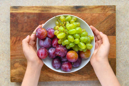 women's hands with a plate full of green grapes and plums on a wooden board, the concept of fresh...