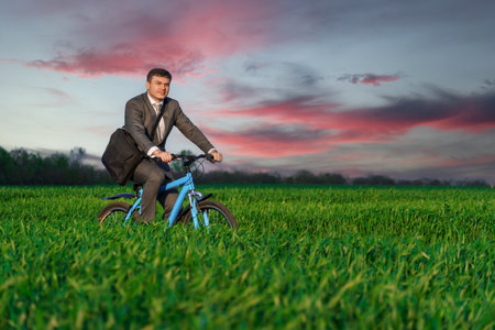 a businessman rides a bicycle with a briefcase on a green grassy field, dressed in a business suit, beautiful nature in spring, freelance business conceptの写真素材