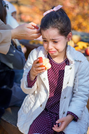The girl is crying and eating an apple, she is sad, she is upset. Mom calms and cares. They are in the autumn city park, a bright sunny day and beautiful nature.の写真素材