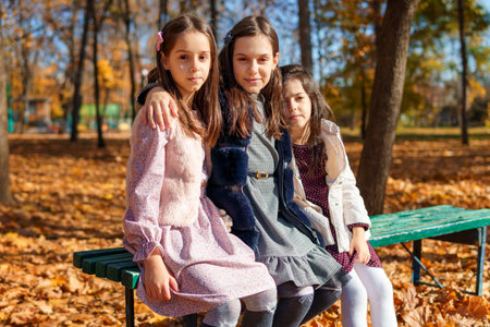 portrait of girls in an autumn city park, children sitting on a bench, talking and enjoying the beautiful nature, talking and playing together, picking yellow leavesの写真素材
