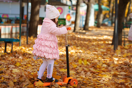 girl rides scooter in autumn city park, she plays, has fun and smiles, yellow maple leaves are around, beautiful nature, bright sunny dayの写真素材