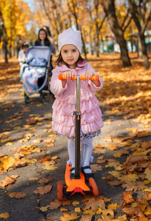 a girl rides a scooter along a path in an autumn city park, a happy child enjoys beautiful nature, a bright sunny day and yellow leavesの写真素材