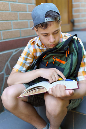 school student, a teenage boy is sitting on the steps near the building, reading a book, a bright summer day, back to school and education conceptの写真素材
