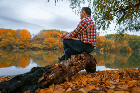 a man sits on a log by the river bank and enjoys the scenery, beautiful nature in the autumn season, the river and the forest with bright yellow leaves, cloudy sky in the eveningの写真素材