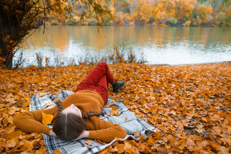a young teenage girl is lying in a glade by the river bank in an autumn forest, enjoying the beautiful nature and bright yellow leavesの写真素材