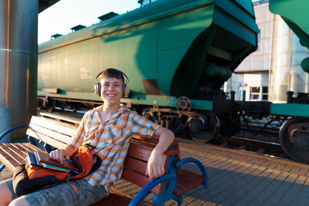 a student poses at a railway station, a boy waits for a train on the platform, listens to music with headphones, reads books and does homework, goes to study, the concept of educationの写真素材