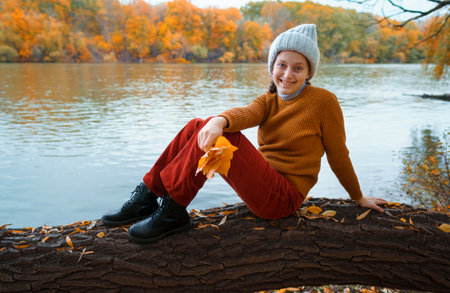 a young teenage girl posing in an autumn forest, sitting on a tree by the river bank, beautiful nature and bright yellow leavesの写真素材