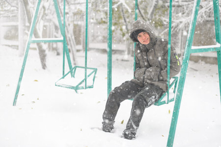 a boy sitting on a swing in the playground, on a snowy street, it is snowing, a blizzard and frost on a winter dayの写真素材