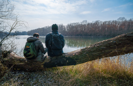 Two men sitting on a log by the river, father and son, early spring landscape, hiking and outdoor activities conceptの写真素材