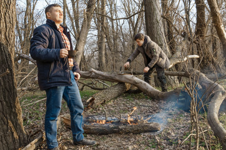 Two men make a campfire for cooking in the forest, father and son sitting on a log while hiking and outdoor activities, early spring landscapeの写真素材