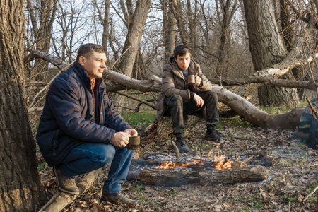 Two men make a campfire for cooking in the forest, father and son sitting on a log while hiking and outdoor activities, early spring landscapeの写真素材