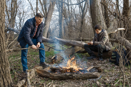 Two men make a campfire for cooking in the forest, father and son sitting on a log while hiking and outdoor activities, early spring landscapeの写真素材