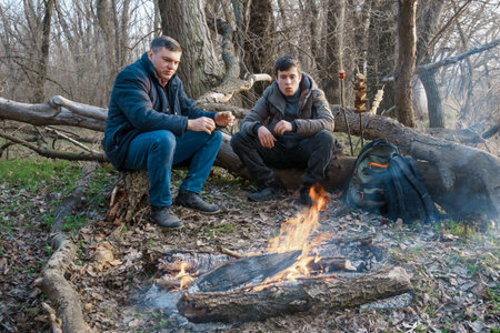 Two men make a campfire for cooking in the forest, father and son sitting on a log while hiking and outdoor activities, early spring landscapeの写真素材