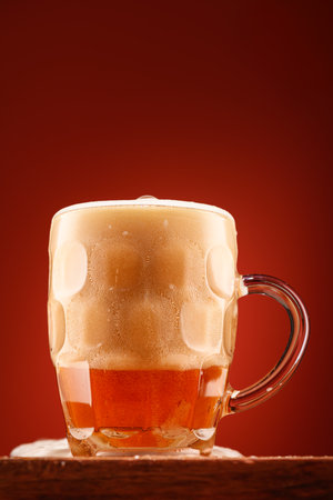 a glass of beer, a wooden stand, a red background, a close-up view of the object, beer bubbles and foamの写真素材