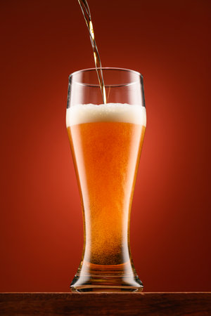 pouring beer into glass, wooden stand, red background, close-up view of object, beer foams and bubblesの写真素材