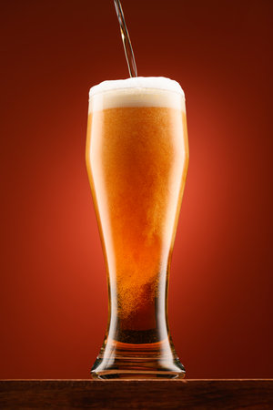 pouring beer into glass, wooden stand, red background, close-up view of object, beer foams and bubblesの写真素材
