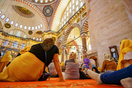 a group of tourists sits on a carpet in mosque and listens to a historical lecture, interior of mosque, domes are painted with patterns, decorative windows and columns, chandeliersの写真素材