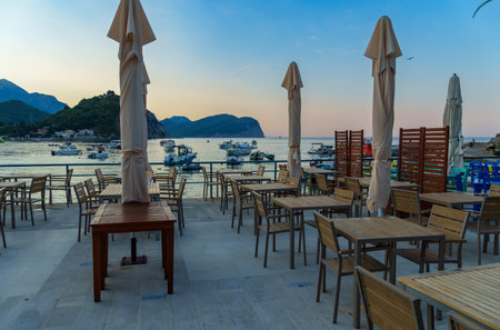 sunrise on adriatic sea coast, street cafe on the embankment of petrovac city in early morning, montenegro, coastline, tables and umbrellas, sun rises over mountainsの写真素材