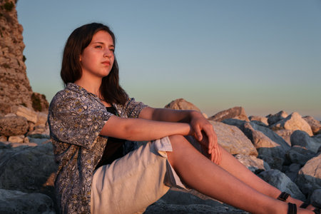 A girl poses and relaxes by the sea, sitting on rocks near the slope of a high cliff, panoramic view of the seashore and mountains at sunsetの写真素材