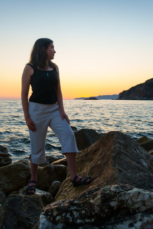 A girl poses and relaxes by the sea, on the rocks, a panoramic view of the coast and mountains at sunset, sunlight creates backlighting and silhouettesの写真素材