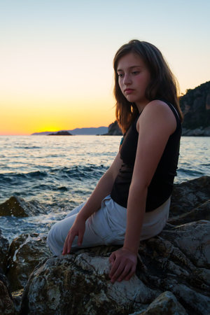 A girl poses and relaxes by the sea, on the rocks, a panoramic view of the coast and mountains at sunset, sunlight creates backlighting and silhouettesの写真素材