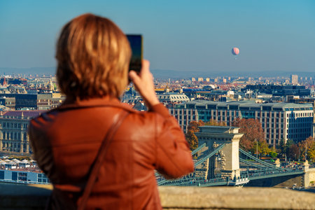 A tourist takes a photo of the Budapest panorama, a beautiful cityscape overlooking the city and the Danube River with the Chain Bridge and beautiful architecture in the autumn season.の写真素材