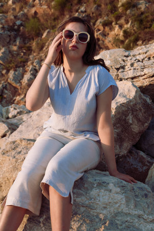 Fashionable young woman adjusting her sunglasses while sitting on the rocky shore during golden hour.の写真素材