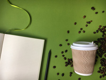 An open lined notebook with a pencil, takeaway coffee cup with a wavy pattern, and scattered coffee beans on a green background, ideal for illustrating study, work, or a coffee break.の写真素材
