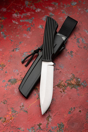 A modern hunting knife with a black handle and sheath on a red, weathered surface.