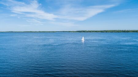 A small sailing riverboat of white color sailing along the sandy riverbank overgrown with green grass. View from above.の写真素材
