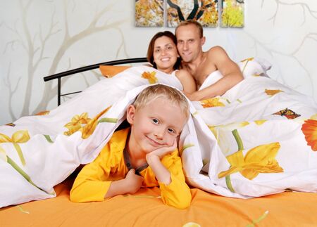 Smiling little boy under the covering in bed with his parentsの写真素材