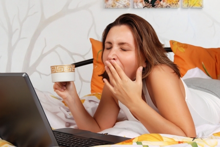 Yawning pretty girl in her bedroom with cup of coffee and notebook in morning timeの写真素材