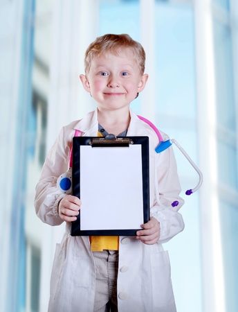 Cute little boy in doctor uniform with clip board in hands on light blue hospital backgroundの写真素材