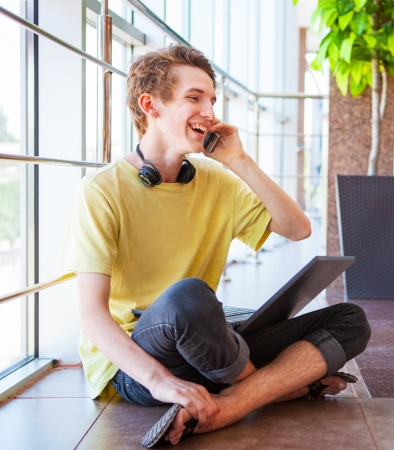 Cheerful smiling teenage boy talking by phone sitting on the floorの写真素材