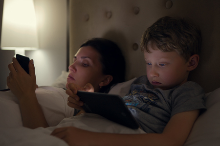 Mother with son looks in their electronic devices lying in bedの写真素材