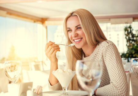 Positive young woman eating ice cream in light summer cafeの写真素材