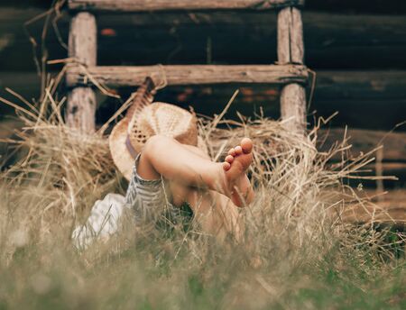 Barefoot boy sleeps on the grass near ladder in haystackの写真素材