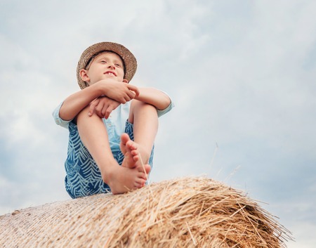 Boy sits on the haystack top with sunny sky backgroundの写真素材